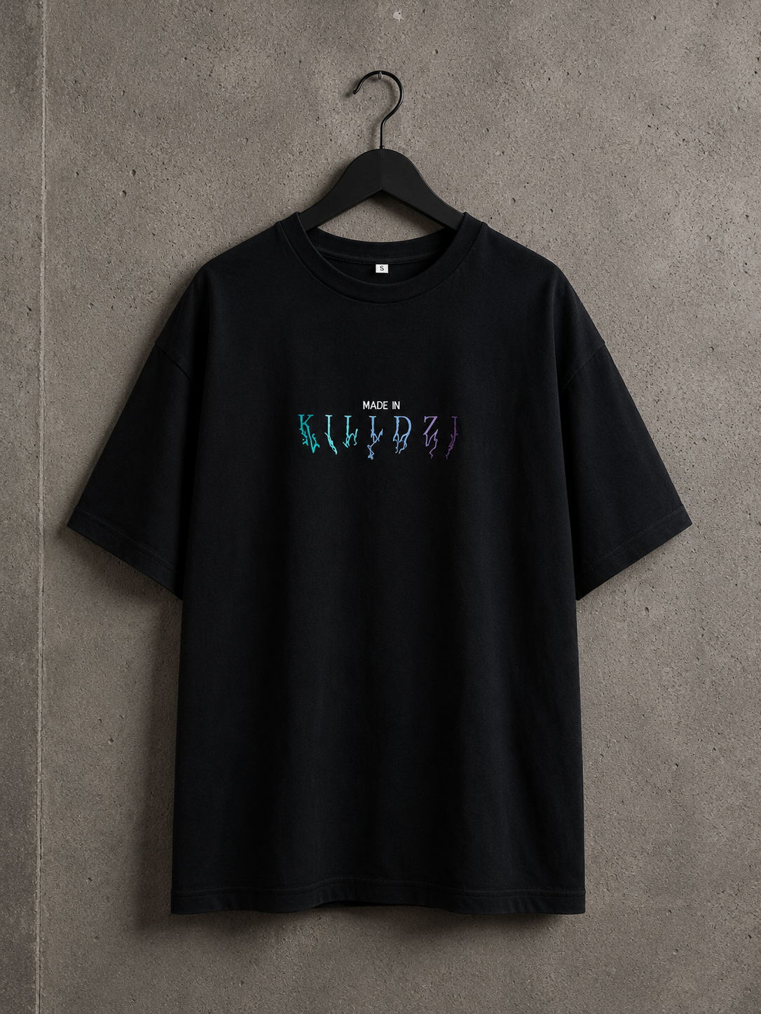 T-shirt oversize noir Made in Killdzi avec logo brodé multicolore, suspendu sur cintre sur fond béton industriel.