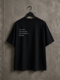 Vue arrière du t-shirt oversize noir Made in Killdzi avec citation brodée à la main, présenté sur cintre devant un mur en béton