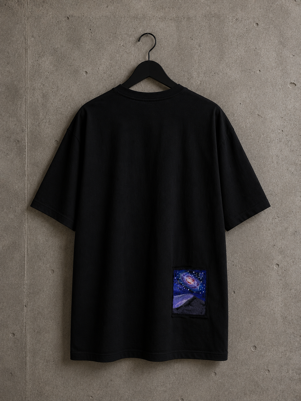 T-shirt oversize Made in Killdzi avec broderie artisanale galaxie au Needle Felting, présentation sur cintre avec fond en béton.