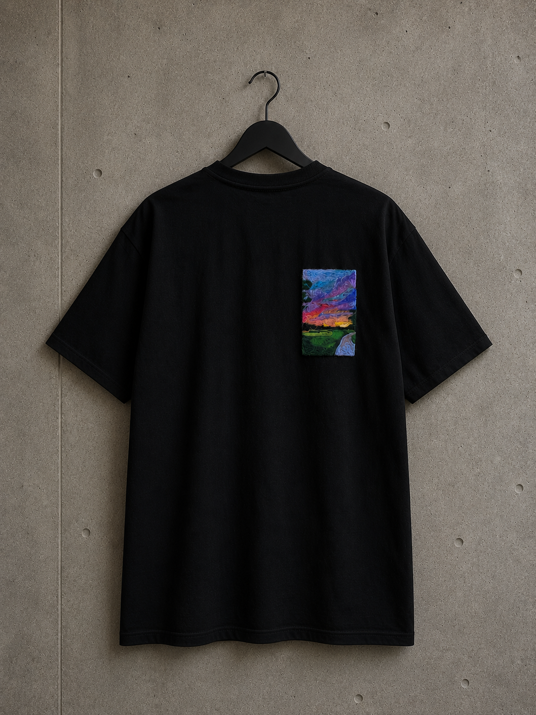 Dos d'un t-shirt oversize noir Made in Killdzi avec broderie artisanale colorée au Needle Felting représentant un paysage de coucher de soleil,, présenté sur cintre sur fond béton